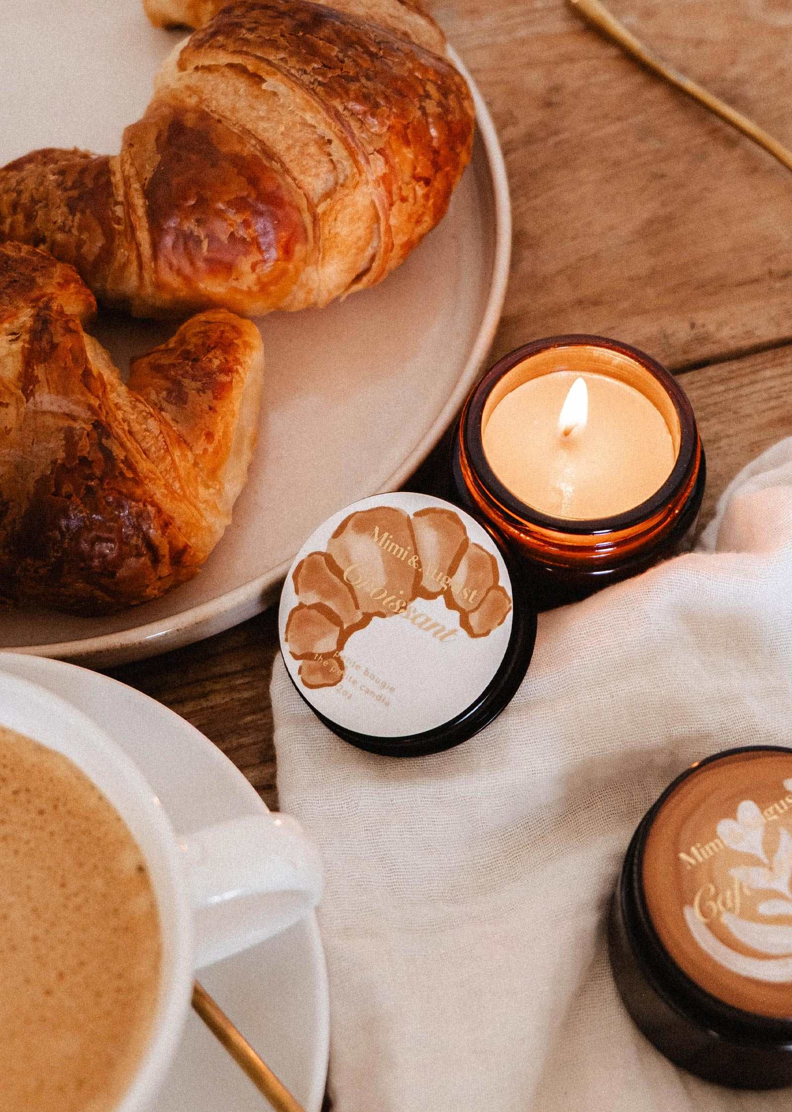 Une surface en bois accueille un croissant, un café et la bougie de soya Croissant de 2 oz de Mimi & August, évoquant le parfum réconfortant d'un matin de boulangerie française.