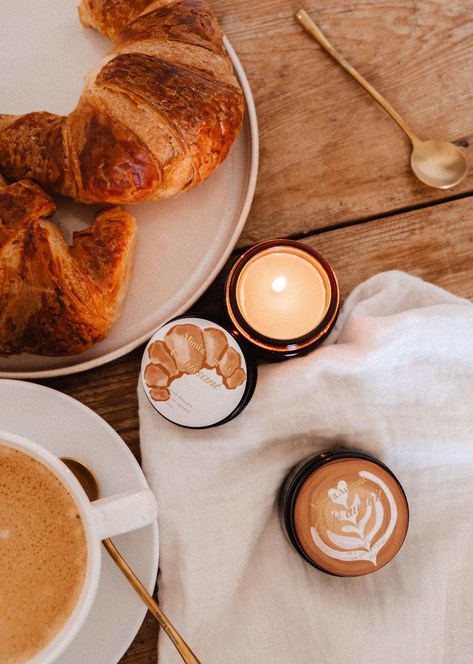 Une bougie de soya Croissant de 2 oz de Mimi & August brille à côté d'un vrai croissant, de deux petits pots, d'un café et d'une cuillère dorée, le tout disposé sur une table en bois avec un tissu blanc, évoquant l'arôme réconfortant d'une boulangerie française.