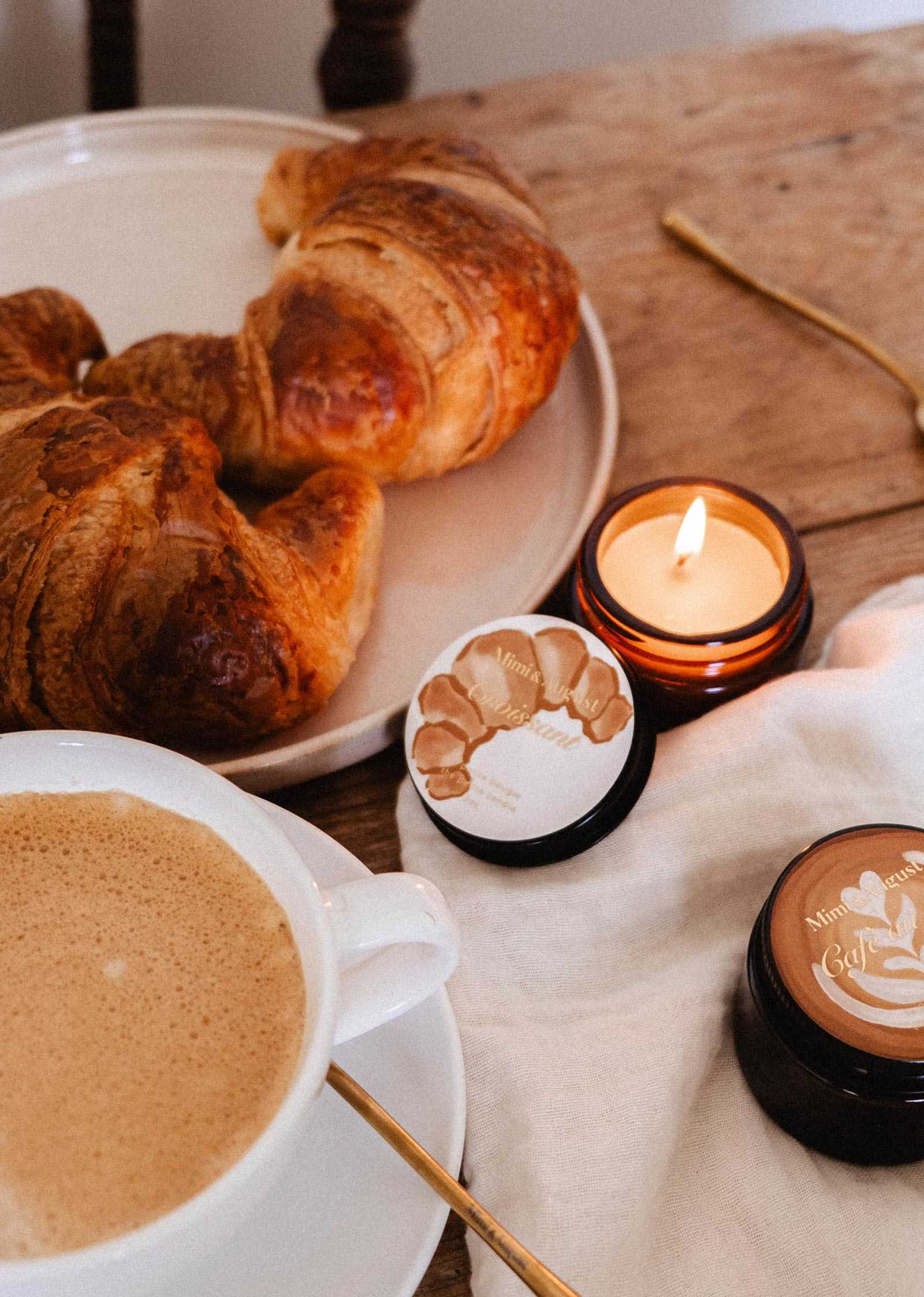 Une bougie de soya Croissant de 2 oz de Mimi & August repose sur une table en bois avec un tissu blanc, son subtil parfum de boulangerie française emplissant l'air à côté de deux croissants, d'une tasse de café et de deux pots de crème pour la peau.