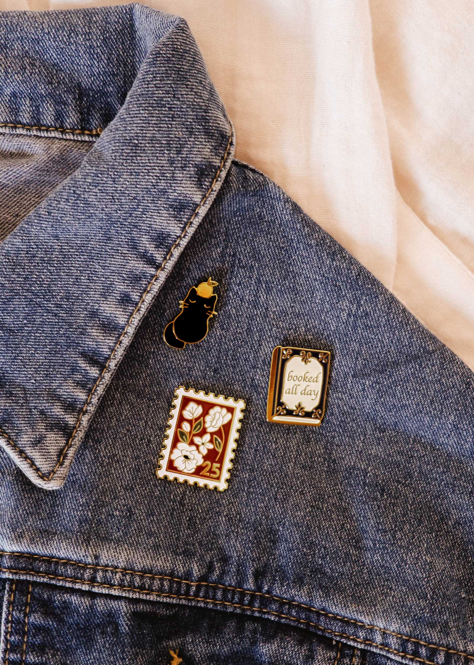 Three Mimi & August enamel pins—a black cat, a "25" floral stamp, and the "Booked All Day" Booked All Day Enamel Pin—adorn a blue denim jacket collar, creating a charming bookworm gift.