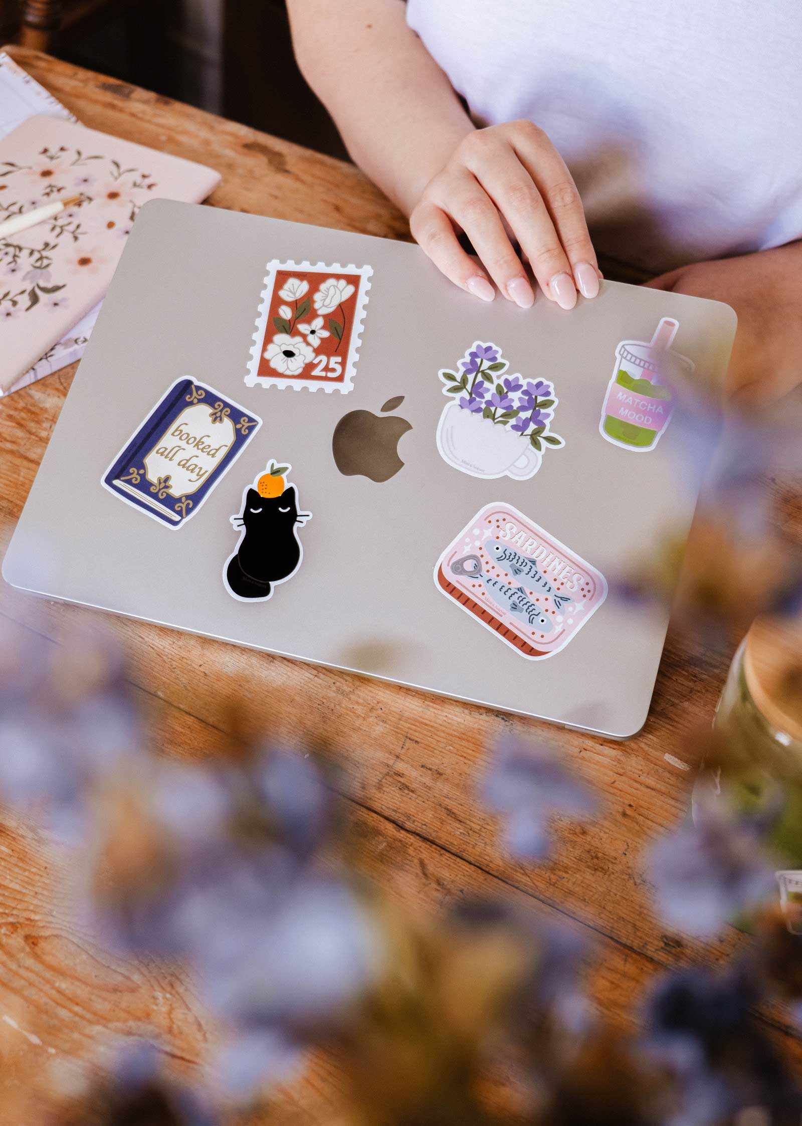 A person rests their hand on a silver laptop adorned with colorful stickers, including the Floral Stamp - Vinyl Sticker by Mimi & August, alongside cat, drink, book designs. A notebook and blurred flowers are nearby.