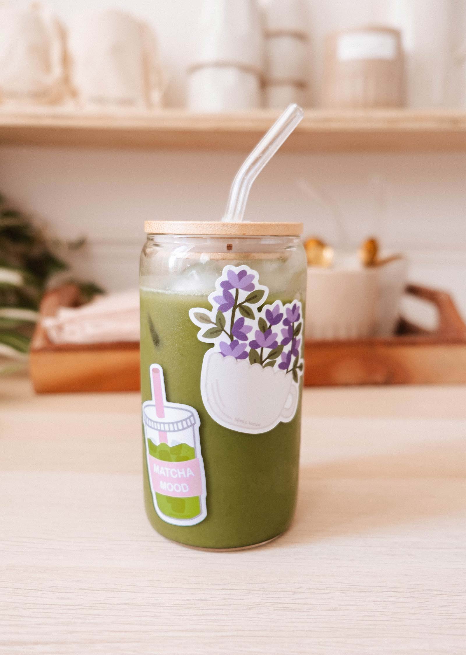 A glass jar of iced matcha latte with a bamboo lid, clear straw, and the Matcha Mood - Vinyl Sticker by Mimi & August sits on a light wooden table, with blurred kitchen items visible in the background.
