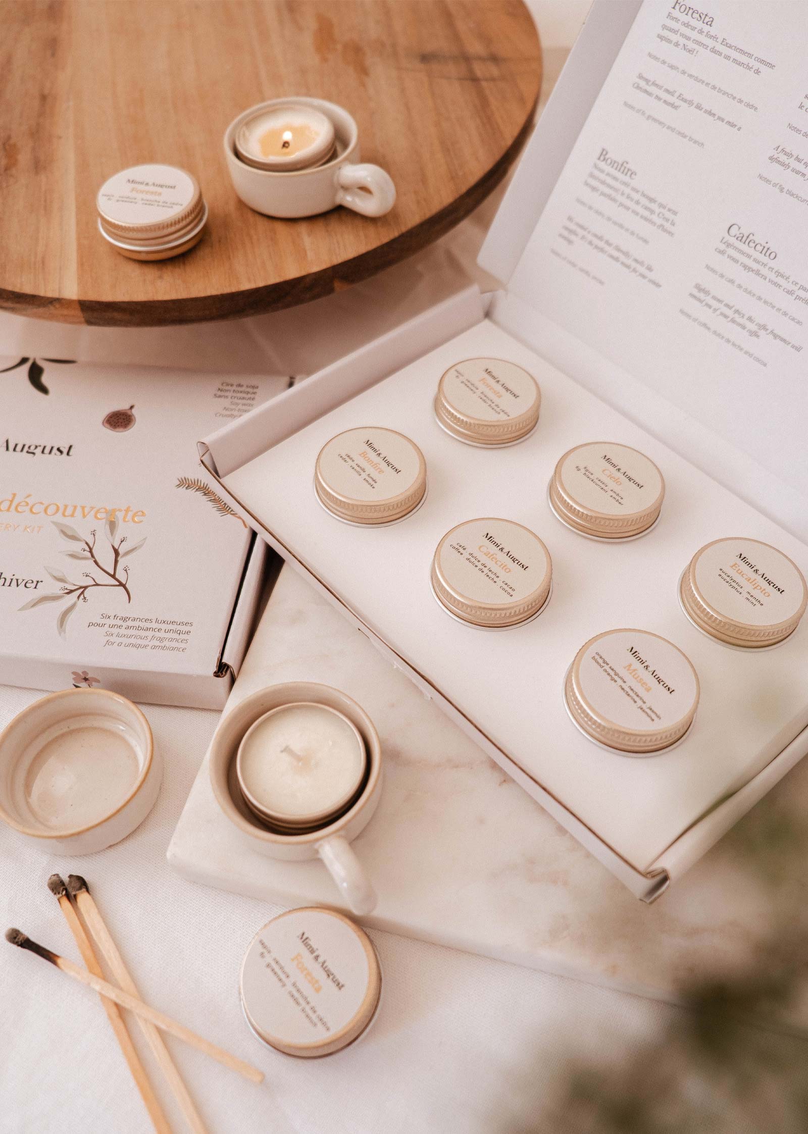 The Fall/Winter Discovery Kit by Mimi & August features small candle tins in an open white box with festive holiday scents, matches, and a lit coffee-scented candle nearby.