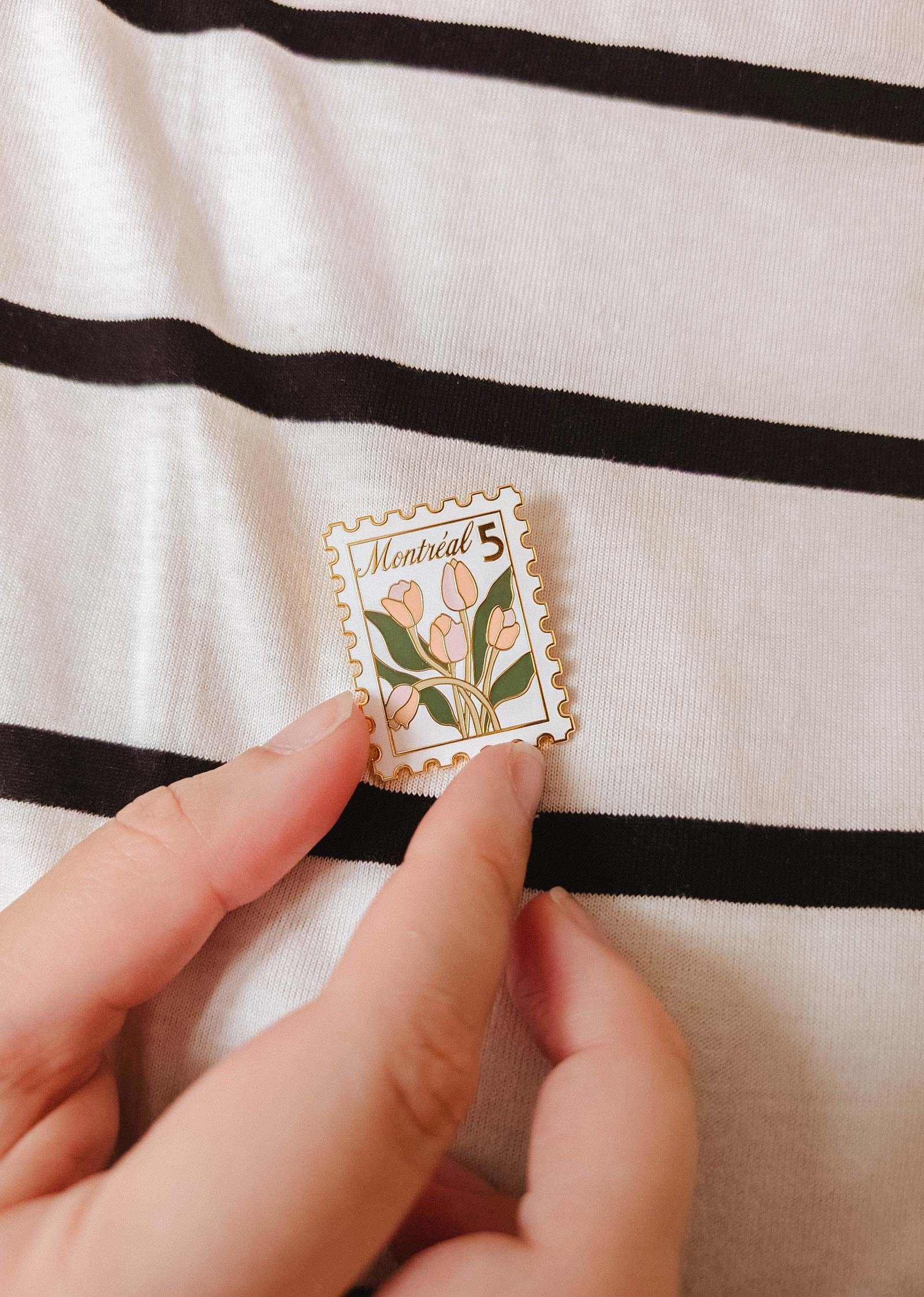 A hand holds the Montréal Stamp Enamel Pin by Mimi & August—an enamel pin styled like a postage stamp with tulips and "Montreal 5" text—set against a black and white striped fabric background. Perfect for flower lovers.