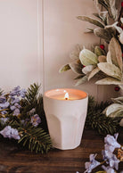 The Mimi & August Special Edition Candle in a ceramic holder is lit, set on wood and surrounded by pine, light purple flowers, green foliage, and red berries.