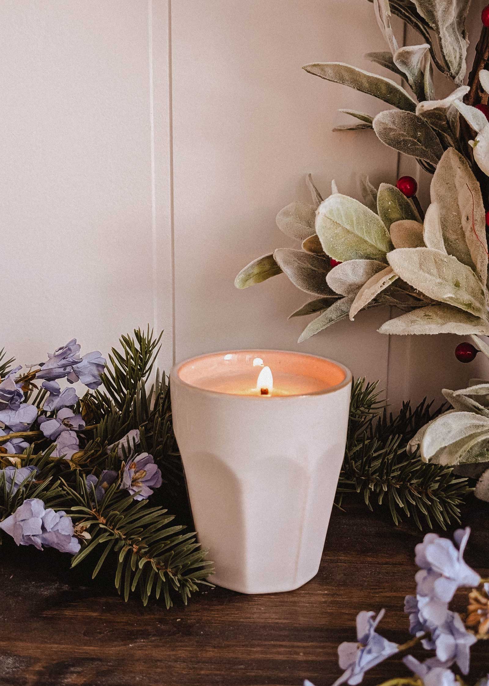 The Mimi & August Special Edition Candle in a ceramic holder is lit, set on wood and surrounded by pine, light purple flowers, green foliage, and red berries.