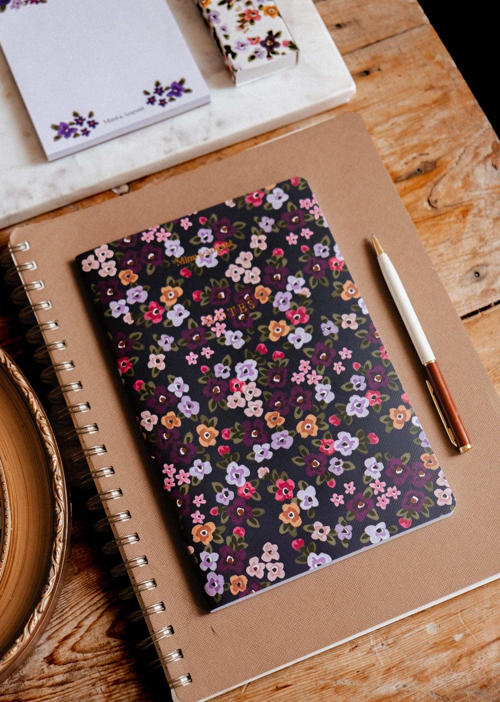 Fields of Flower floral notebook by Mimi & August on a wooden table with a pen, sketchbook, and matching notepad.