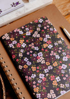 Fields of Flower floral notebook by Mimi & August resting on a brown spiral notebook beside a white pen.