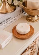 A white, fluted-edge Candle Refill Topaze by Mimi & August sits on a wooden dish near stacked books, a gold bowl, and a "Mini Acust" box, filling the space with sandalwood coconut vanilla warmth.