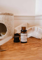 A brown bottle of fig scented oil - Cielo by Mimi & August with a black cap, positioned on a wooden surface beside a ceramic diffuser. The bottle's label reads "Mimi & August Cielo." The cap is off and resting next to it, allowing a calming scent to fill the air.