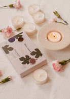 A lit fig tealight candles on a ceramic holder amidst pink flowers creates a cozy ambiance. The Cielo Tealight Candle Set from Mimi & August, promising a warm comforting scent, sits gracefully on the white surface.