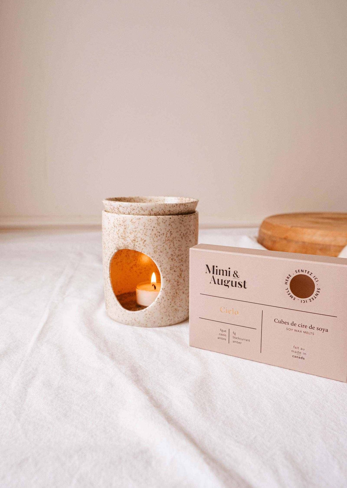 A ceramic candle holder with a lit tealight sits next to a box of Mimi & August's fig wax melts - Cielo on a white cloth, promising a long-lasting fragrance.