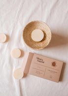 A speckled bowl cradles a cream-colored wax melt made from 100% soy. An open "fig wax melts - Cielo" package by Mimi & August promises lasting fragrance, with extra melts laid on a white fabric.