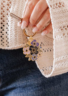 A hand holds the Mimi & August Floral Bouquet Keychain, showcasing purple and black enamel flowers, paired with a cream knit sweater and blue jeans. This Montreal-designed accessory adds a charming touch to any outfit.