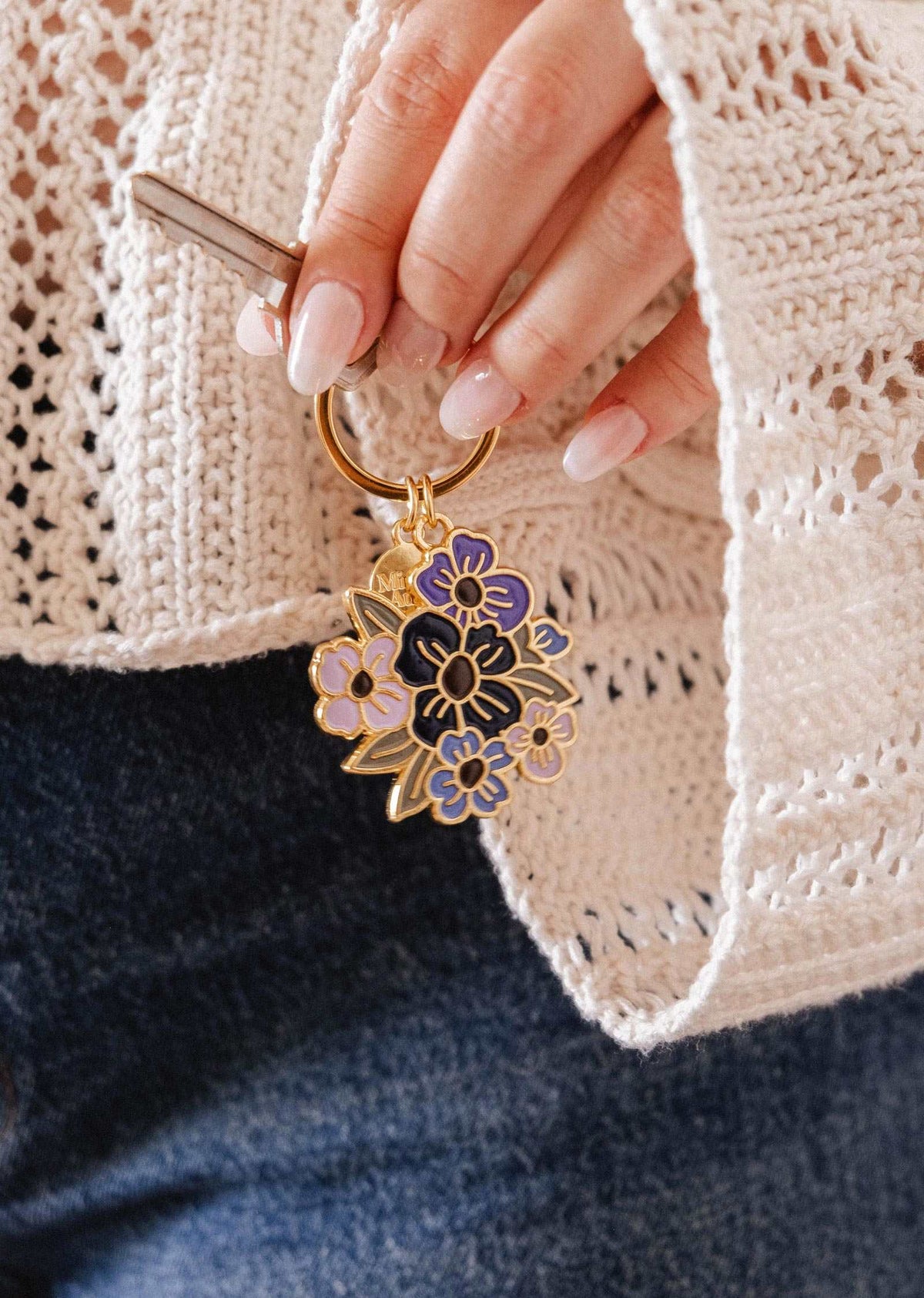 A hand holds the Mimi & August Floral Bouquet Keychain, showcasing purple and black enamel flowers, paired with a cream knit sweater and blue jeans. This Montreal-designed accessory adds a charming touch to any outfit.