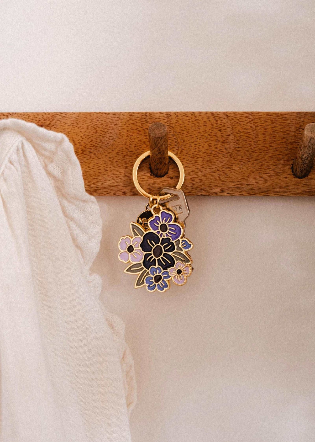 A gold Floral Bouquet Keychain from Mimi & August, featuring a purple and black enamel design, hangs on a wooden hook beside a ruffled white fabric—a charming accessory designed in Montreal.