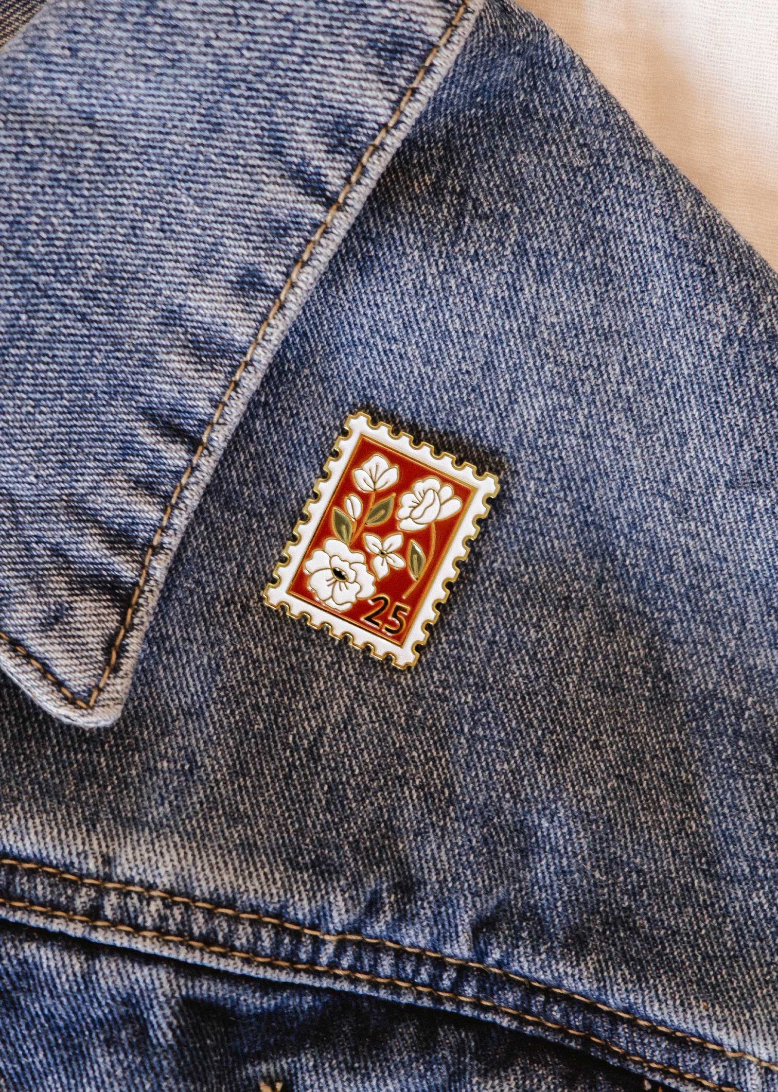 The Mimi & August Floral Stamp Enamel Pin, shaped like a vintage stamp with a floral charm and the number 25, is attached to a blue denim jacket.