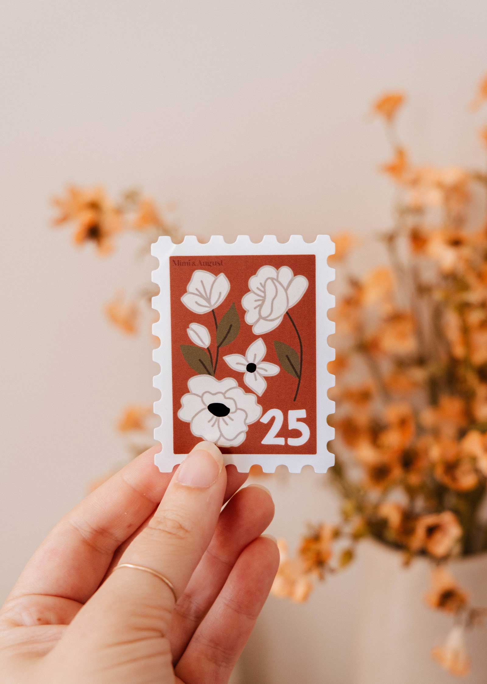 A hand holds the Mimi & August Floral Stamp - Vinyl Sticker, featuring white flowers and the number 25, with blurred dried flowers in the background.
