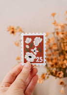 A hand holds the Mimi & August Floral Stamp - Vinyl Sticker, featuring white flowers and the number 25, with blurred dried flowers in the background.