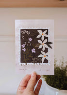 A hand holds the "Dimanche Fleuri - Free Art Print (100% off)" by Mimi & August, featuring white and purple flowers with green leaves in a stamp-like frame—perfect as a free gift from BOGOS.io.