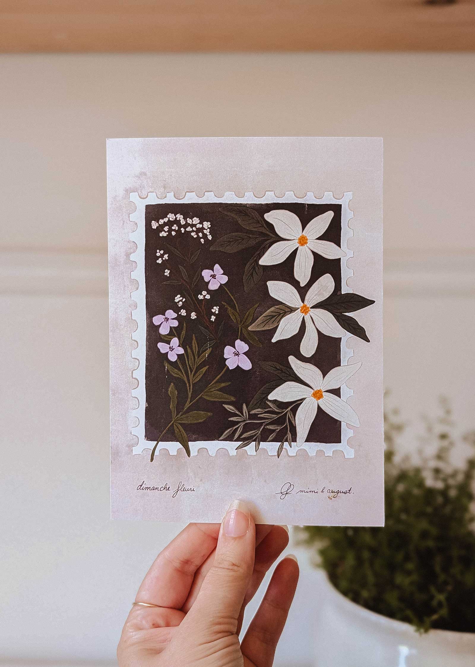 A hand holds the "Dimanche Fleuri - Free Art Print (100% off)" by Mimi & August, featuring white and purple flowers with green leaves in a stamp-like frame—perfect as a free gift from BOGOS.io.