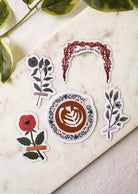 The Floral Love – Free Sticker Set (5 pcs) by Mimi & August features flowers, latte art, and a red floral arch—arranged on marble with green leaves visible. Get it 100% off as a Free Gift from BOGOS.io or add to your BOGO Bundle!.