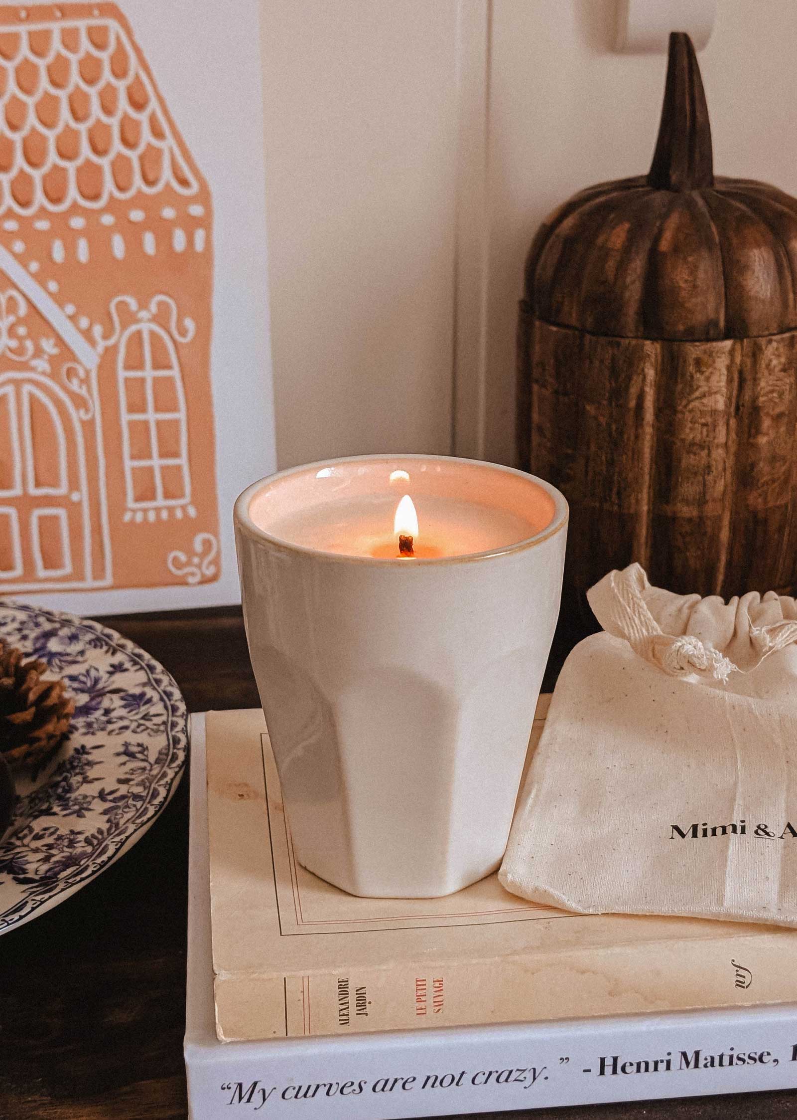The Special Edition Candle by Mimi & August in a white ceramic holder rests on a stack of books, accompanied by a small drawstring bag and decorative items.