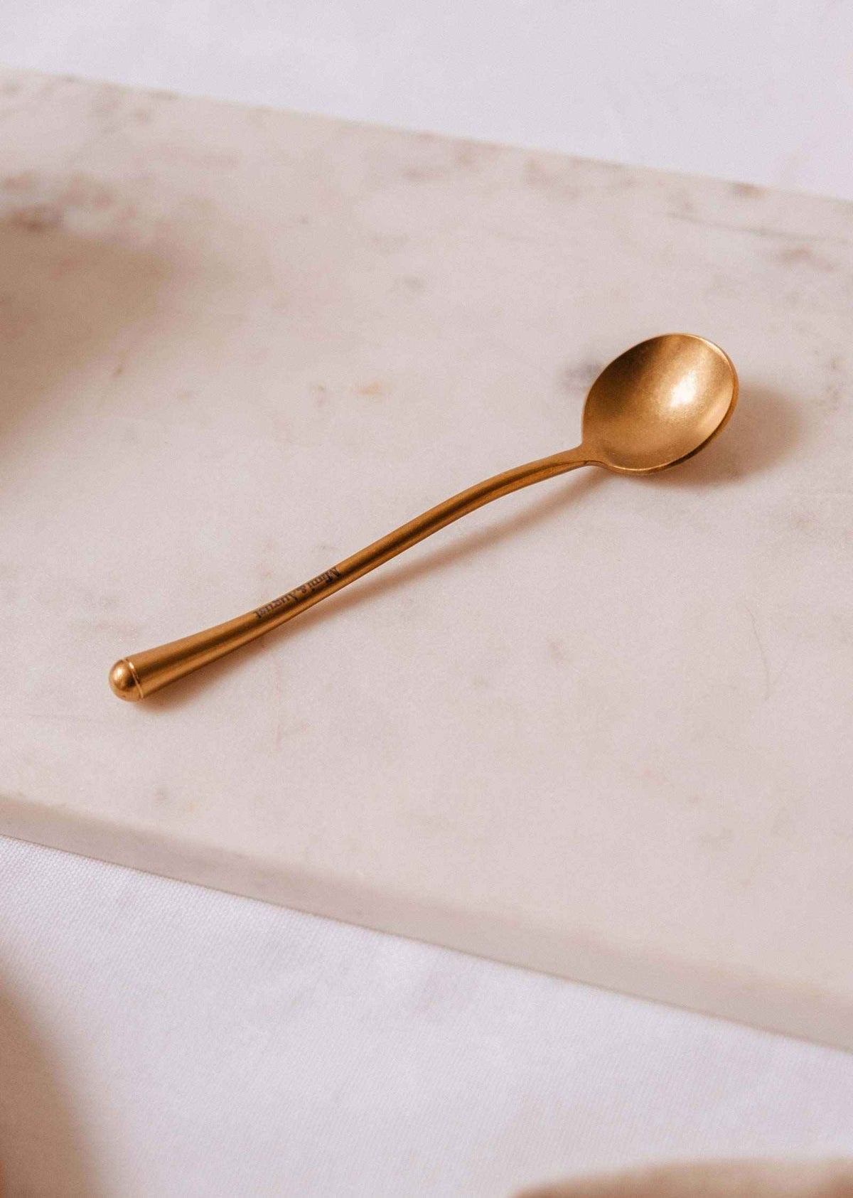 The Golden Touch – Delicate Gold Teaspoon by Mimi & August shines atop a white marble surface.