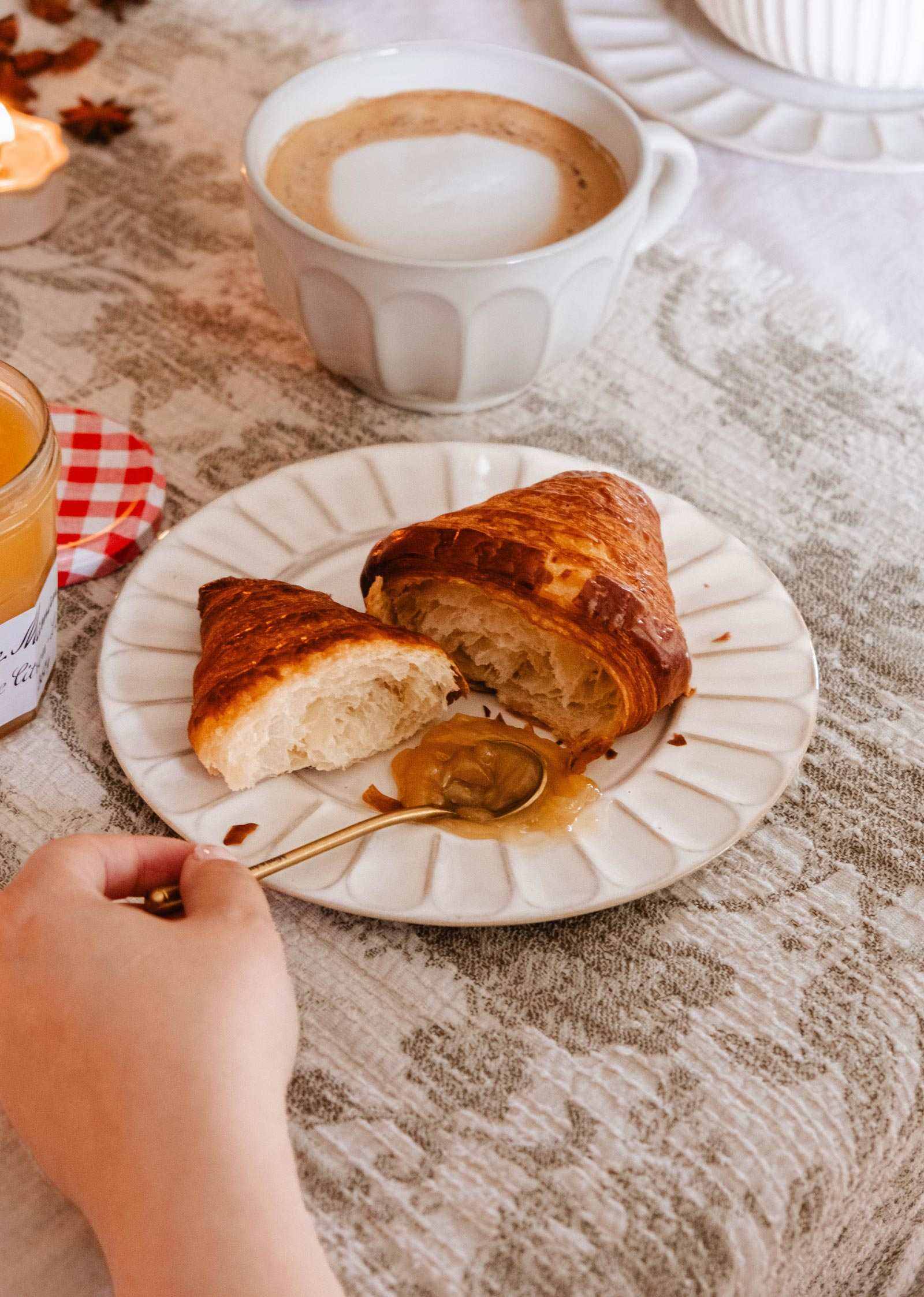 A hand holds a spoon with honey above handmade ceramic plate by Mimi & August, which displays a sliced croissant beside a cup of coffee on a table draped with a white tablecloth.