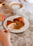 A hand holds a spoon with honey above handmade ceramic plate by Mimi & August, which displays a sliced croissant beside a cup of coffee on a table draped with a white tablecloth.