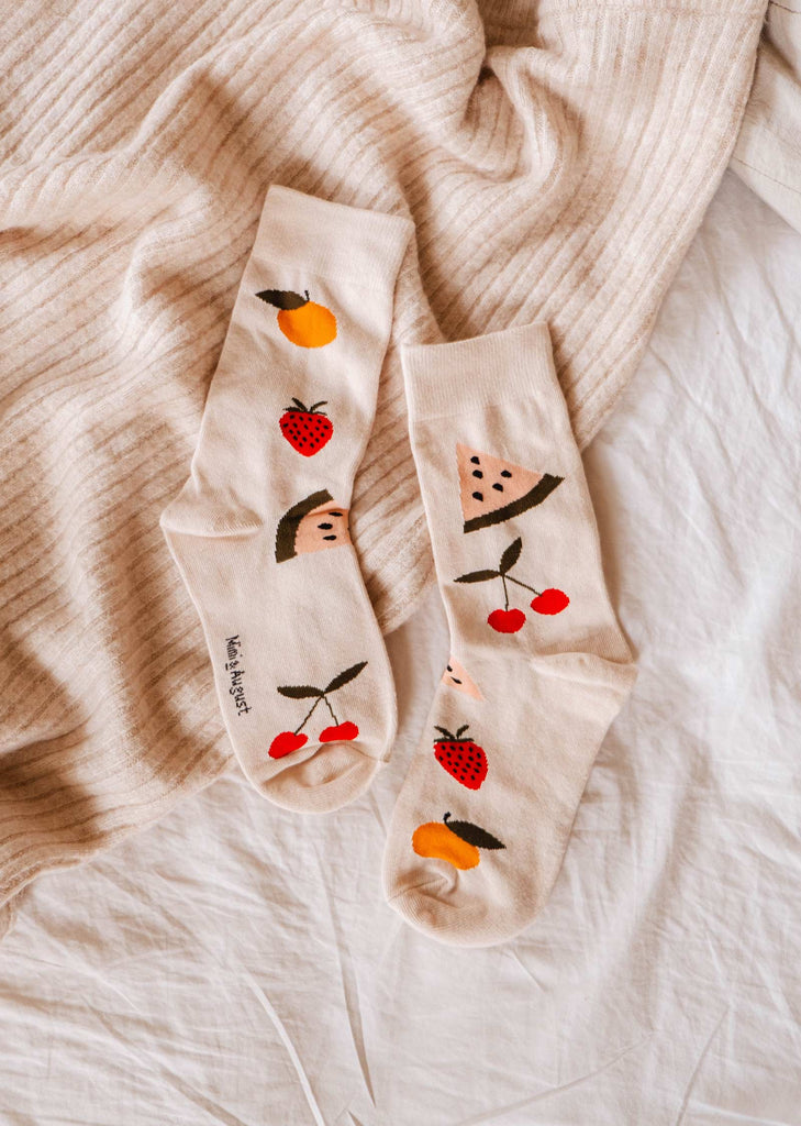 Deux paires de chaussettes Happy Fruits de Mimi & August, de couleur beige, représentant des illustrations de fruits ludiques tels que des fraises, des cerises et des oranges, sur un tissu côtelé beige et un fond blanc. Idéales pour être portées tous les jours, ces chaussettes confortables et fantaisistes sont un ajout charmant à votre garde-robe.