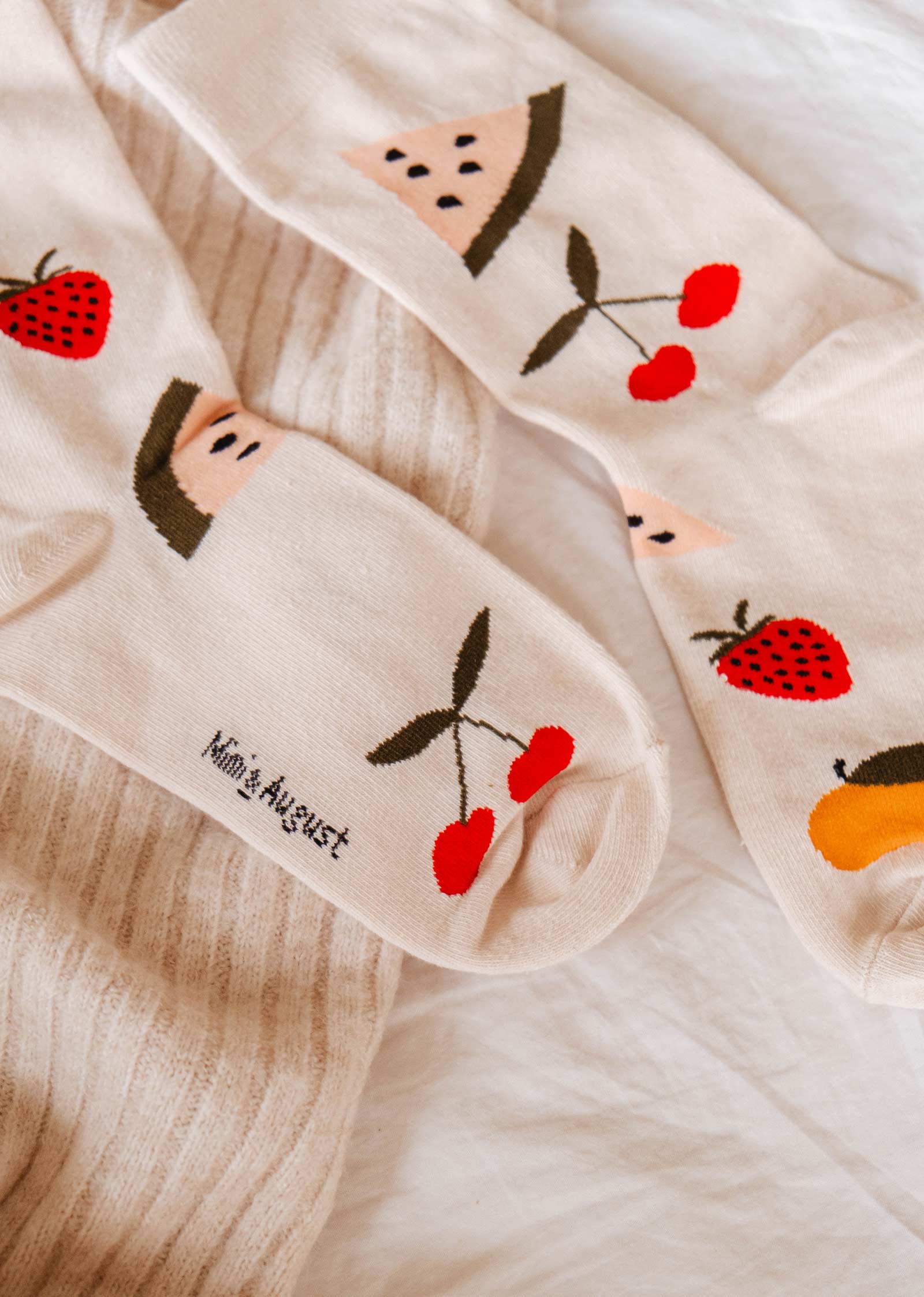 A pair of comfortable Happy Fruits Socks by Mimi & August adorned with playful fruit patterns including watermelon, strawberries, cherries, and peaches, lying on a cream-colored fabric. Perfect for everyday wear.