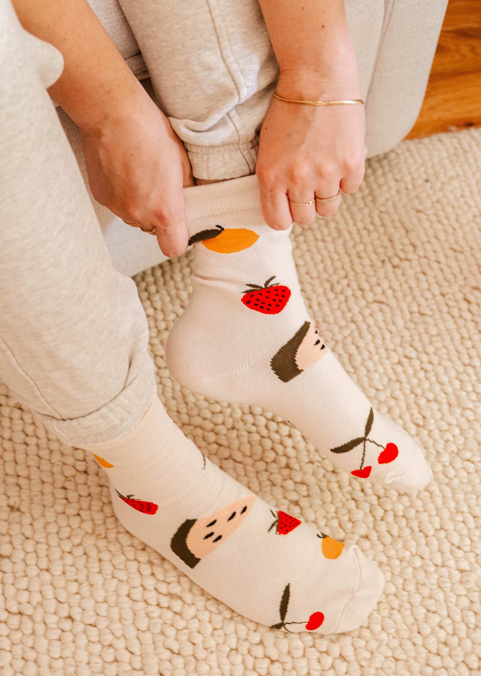 A person is putting on comfortable Happy Fruits Socks by Mimi & August, which are decorated with fruits and faces, while wearing light grey pants and seated on a beige carpet.