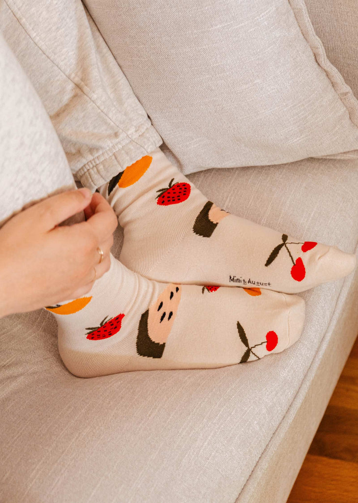 Une personne portant des chaussettes Happy Fruits de Mimi & August avec des motifs de fruits, assise sur un canapé et ajustant ses chaussettes confortables. Les motifs comprennent des cerises, des fraises, des oranges et des pastèques. Parfaites pour la vie de tous les jours.