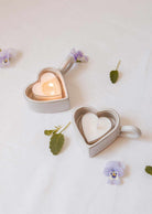 Deux Bougeoirs Cœur & Lueur de Mimi & August, l'un avec un lampion allumé et l'autre éteint, reposent sur un tissu blanc décoré de fleurs violettes éparpillées et de feuilles vertes.
