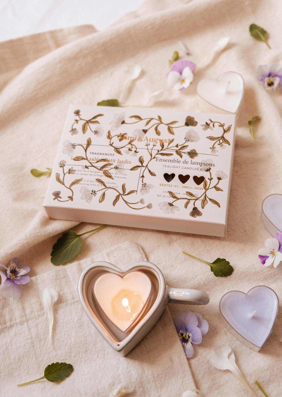Un Bougeoir Cœur & Lueur de Mimi & August repose sur un tissu beige, entouré de lampions Blush Hearts, de bougies en forme de cœur, de pétales de fleurs et de verdure.