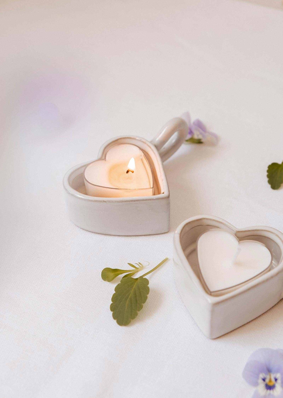 Deux Bougeoirs Cœur & Lueur de Mimi & August, l'un allumé et l'autre éteint, reposent sur une surface blanche avec des fleurs délicates et des feuilles éparpillées à proximité, mettant en valeur leur design en céramique en forme de cœur.
