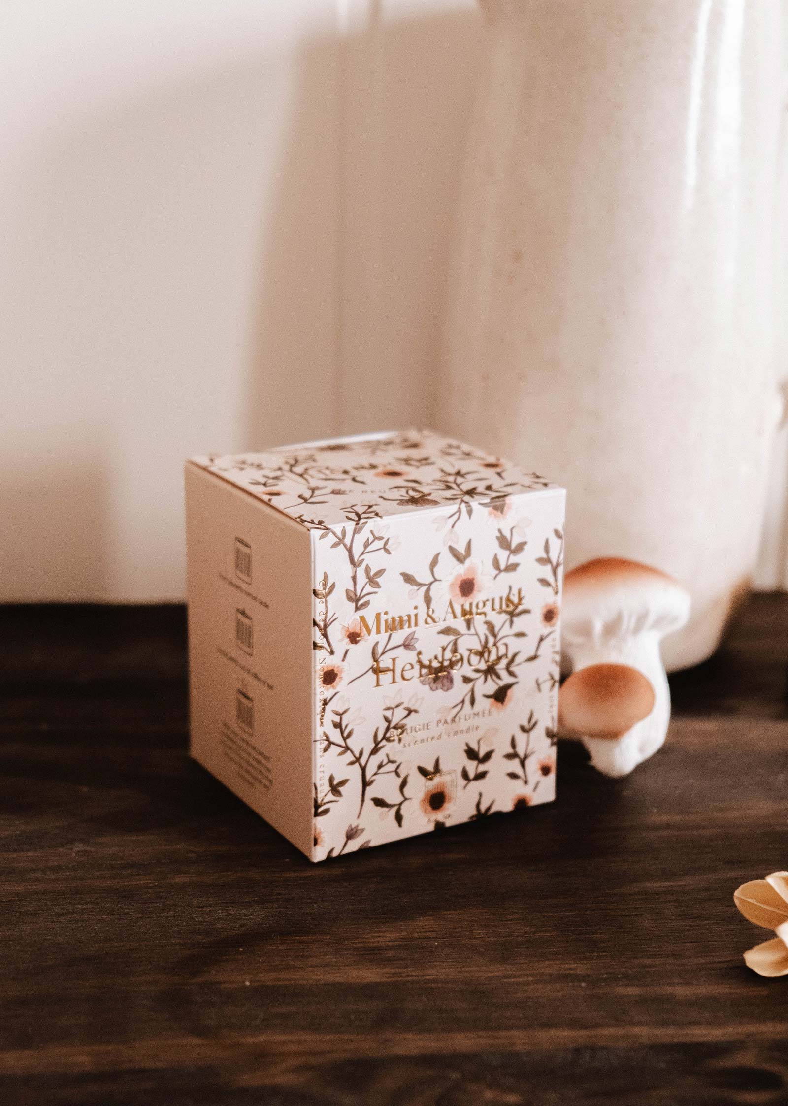 The Mimi & August Heirloom - Reusable Candle, in a decorative box with floral patterns and gold text, sits on a dark wooden surface beside a ceramic vase and two small mushroom ornaments.