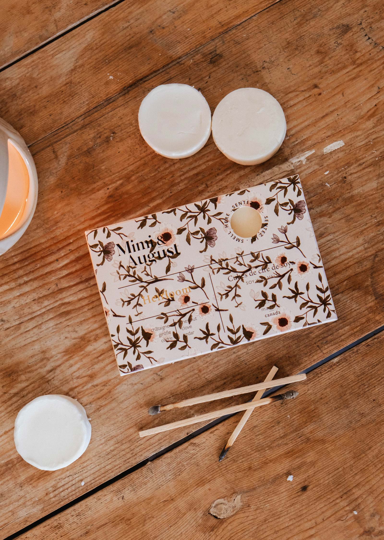 A decorative "Mimi & August" box labeled "Soy Wax Melts - Heirloom" rests on wood, surrounded by three white round melts and four wooden sticks—two with burnt tips—evoking the cozy charm of natural home fragrance.