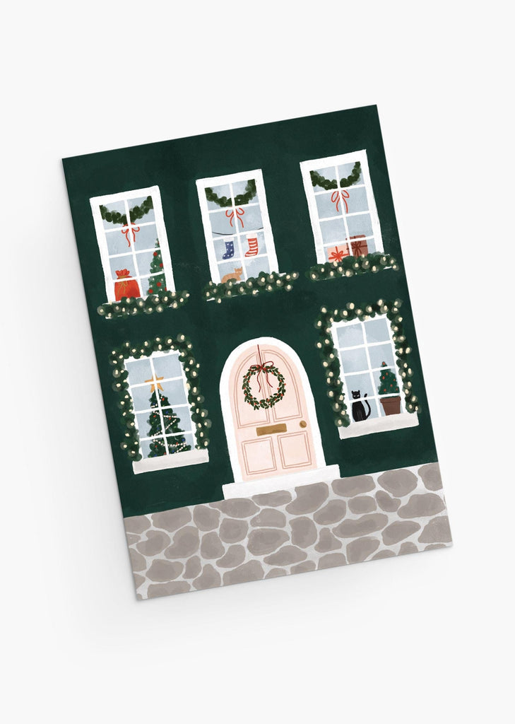 Mignonne carte de vœux de Noël représentant une maison avec une couronne de Noël à la porte, et des chatons, des chaussettes et des arbres à travers les fenêtres. Par Mimi & August