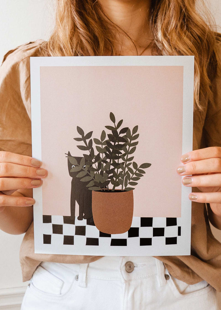 A woman holding up a i see you art print by mimi and august with a cat and a potted plant.