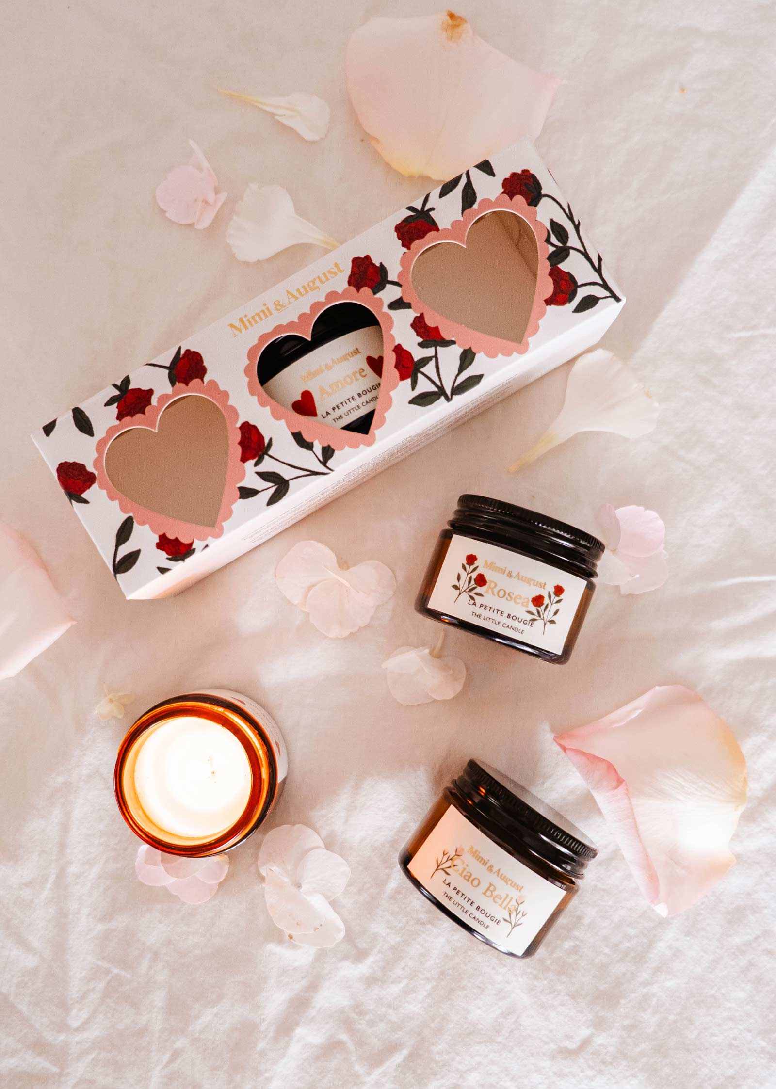 Skincare items, including two jars and an elegant candle, rest on soft pink fabric with scattered petals next to the Mimi & August La Romance Box Set featuring heart-shaped cutouts. With exclusive fragrances, this collection adds luxury to your beauty routine.