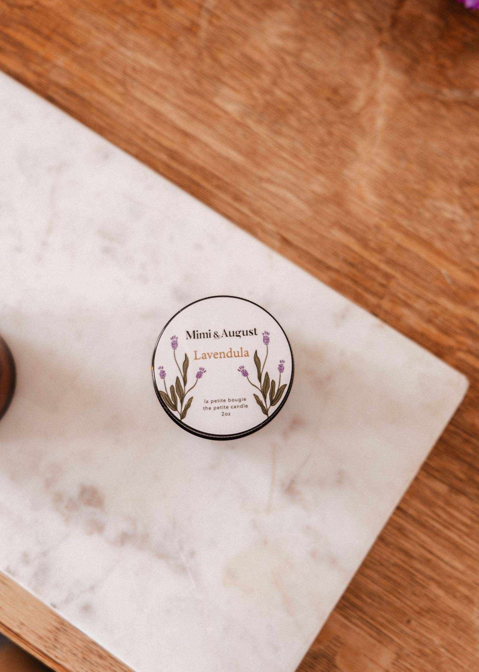 A 2 oz Mimi & August Lavandula soy candle sits on a white marble surface, atop a wooden table.