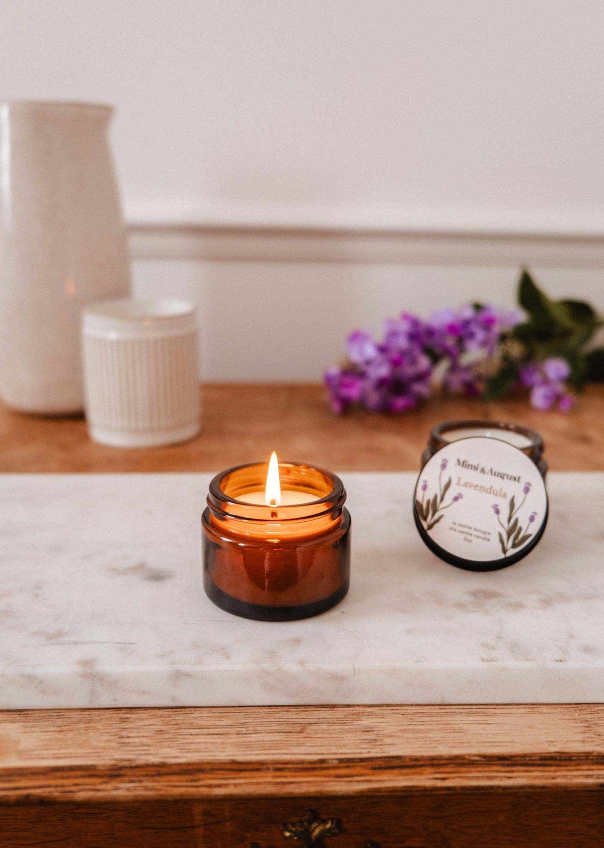 A lit Mimi & August Lavandula – 2 oz Soy Candle sits on a marble tray with purple flowers, a white vase, and a jar in the background, creating a calming candle display.