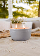 A Glow Candle by Mimi & August flickers softly in its ceramic holder on a textured surface, with blurred outdoor greenery framing the scene. A nearby small plant enhances the serene ambiance.