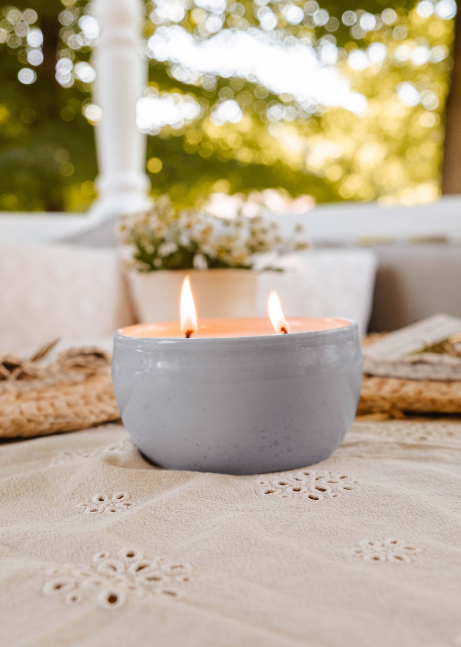 A Glow Candle by Mimi & August flickers softly in its ceramic holder on a textured surface, with blurred outdoor greenery framing the scene. A nearby small plant enhances the serene ambiance.