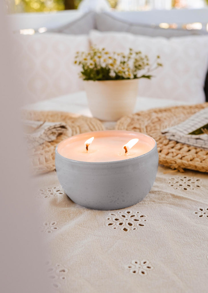 A lit double-wick Bloom Candle from Mimi & August in a handmade ceramic bowl graces the table with its gentle glow, set on a textured tablecloth. In the background, a pot of flowers and textured placemats add to the serene ambiance.