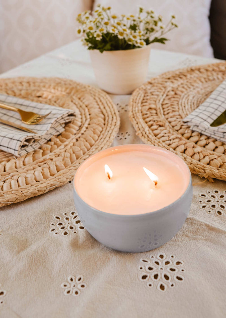 A lit "The Bloom Candle" by Mimi & August on a white table with a lacy cloth, two circular woven placemats, checkered pattern napkins, and a small potted plant in the background.