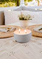 A Glow Candle by Mimi & August in a gray container sits on a table with a floral tablecloth, complemented by a potted plant and woven placemats with cutlery, creating an inviting atmosphere.