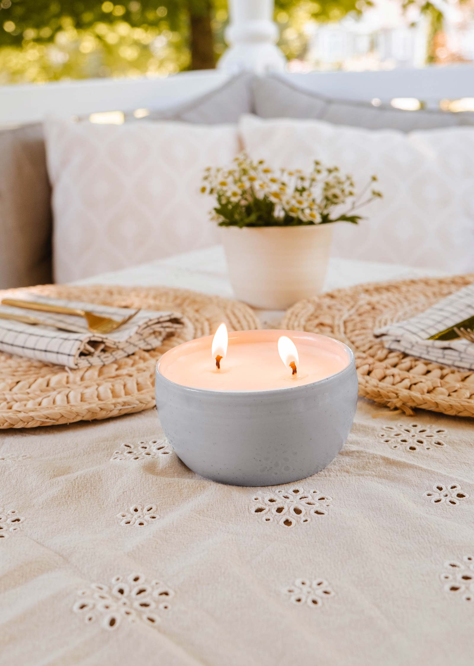 Une bougie Glow Candle de Mimi & August dans un récipient gris est posée sur une table avec une nappe fleurie, complétée par une plante en pot et des sets de table tressés avec des couverts, créant ainsi une atmosphère accueillante.